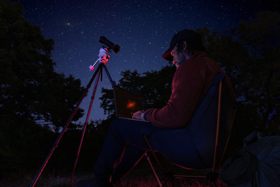 Pessoa usando MacBook Pro M5 do lado de fora à noite com telescópio ao fundo